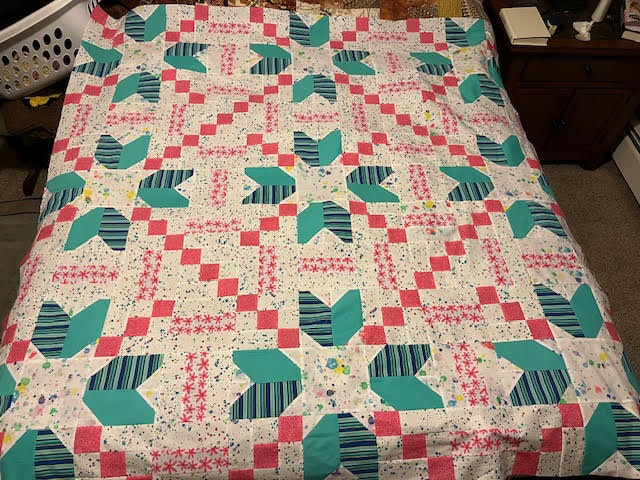 A colorful quilt covers a bed, featuring geometric patterns with stars and arrows. The quilt has a palette of turquoise, pink, and various striped and polka dot designs on a white background.
