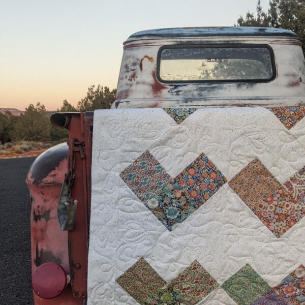 Heart Quilt Pattern 6 A rusted vintage truck is parked on a paved road, with a heart-patterned quilt adorning its tailgate. The sky is dim with a visible moon, while trees stand silhouetted in the background.