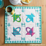 A charming square quilted mat showcases four arrow patterns in blue and pink plaid, evoking a cozy Easter vibe. Each arrow rests on a white background bordered by blue plaid. Nearby, a teapot, cup of tea, and pastries enhance the welcoming scene on the wooden table.
