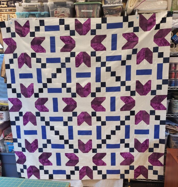 A quilt hangs on a wall, featuring a geometric pattern in purple, blue, and white. The design includes star and cross shapes, with alternating squares creating a symmetrical arrangement. Background storage holds various crafting supplies.