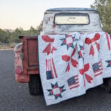 A vintage, rusty pickup truck is parked on a paved road, with a Patriotic Flag Quilt draped over its tailgate. Trees and bushes are visible in the background under a clear sky.