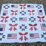 A quilt laid out on gravel, featuring a patriotic flag pattern with red, white, and blue stars and bows, arranged in a grid with a white border. This ribbon quilt has a festive patriotic theme.