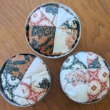 Three round fabric pincushions with patchwork designs in earthy tones and floral patterns, each nestled in a vintage tin. Perfect for DIY enthusiasts, these charming pincushions add a touch of nostalgia to any sewing space.