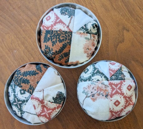 Three round fabric pincushions with patchwork designs in earthy tones and floral patterns, each nestled in a vintage tin. Perfect for DIY enthusiasts, these charming pincushions add a touch of nostalgia to any sewing space.