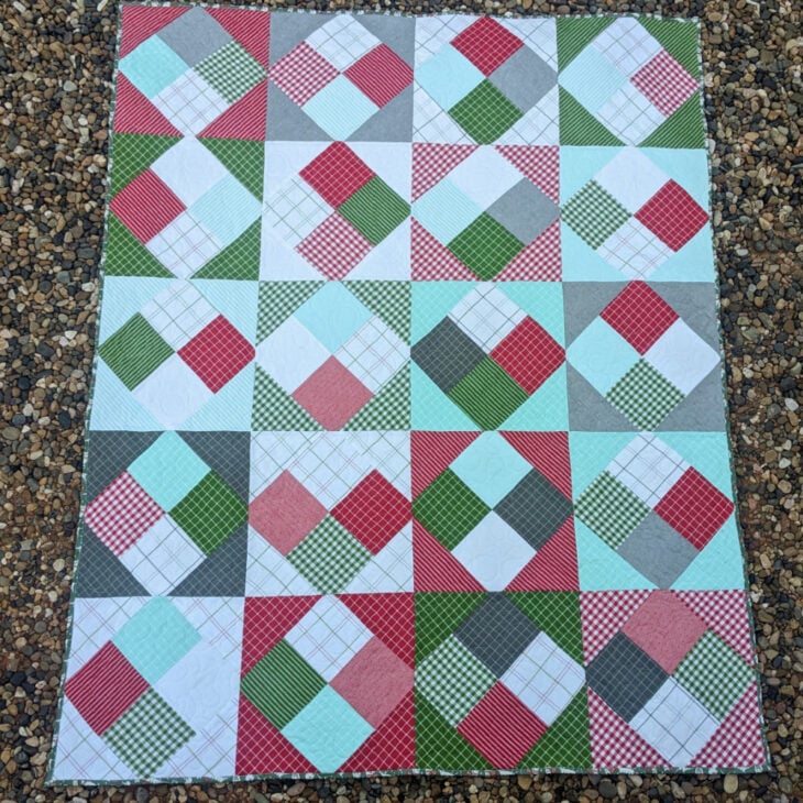 A quilt laid out flat on gravel. the quilt block is a four patch and is on point. the fabric features red, green and white fabric. it's a simple quilt to sew