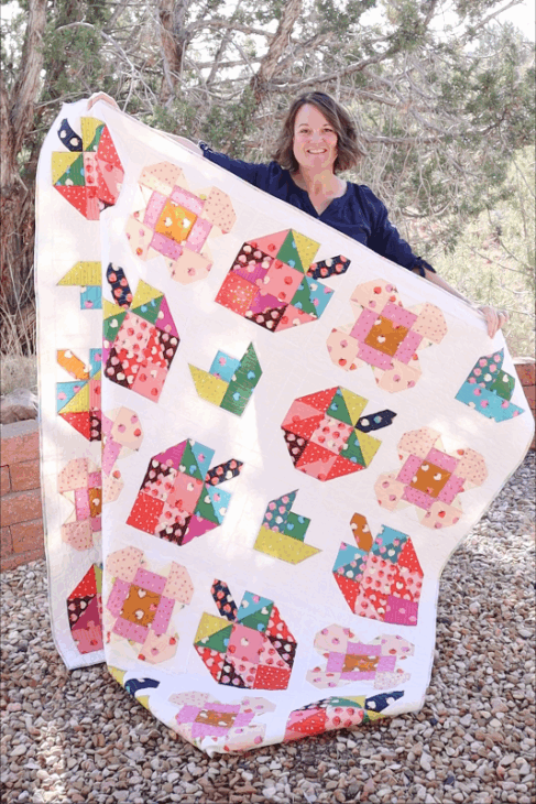 A woman stands outdoors on gravel, smiling and holding up a large quilt decorated with colorful patchwork patterns featuring cats and fish&mdash;an inspiring creation for quilters and any Patchwork Planner enthusiast. Trees and a brick wall are in the background.