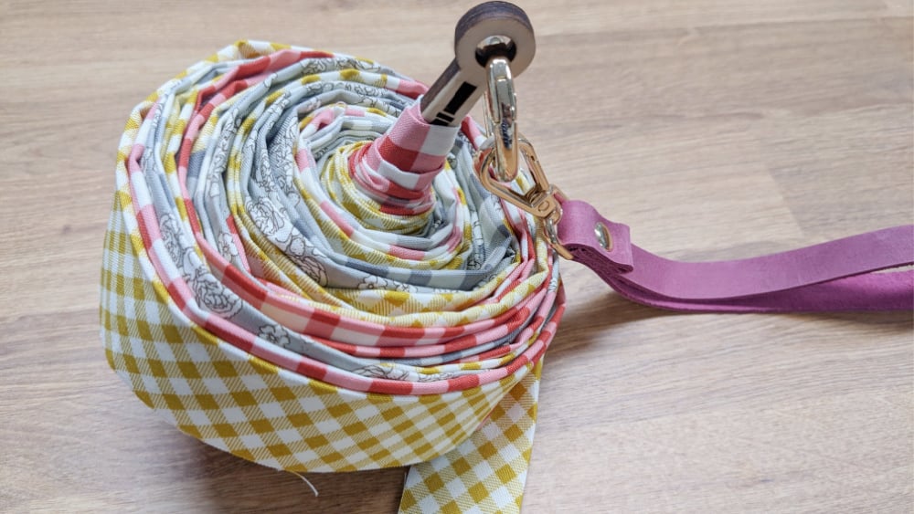 A long, multicolored fabric dog leash with a checkered pattern is coiled neatly on a wooden floor, with a metal clip and a purple handle visible on top.