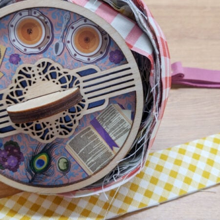 A circular wooden box with decorative patterns and paper cutouts sits on a wooden surface, surrounded by colorful checkered ribbon, including a yellow-and-white strip unfurled to the right.
