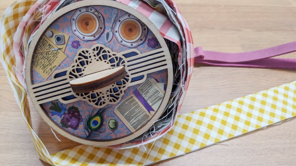 A circular wooden box with decorative patterns and paper cutouts sits on a wooden surface, surrounded by colorful checkered ribbon, including a yellow-and-white strip unfurled to the right.