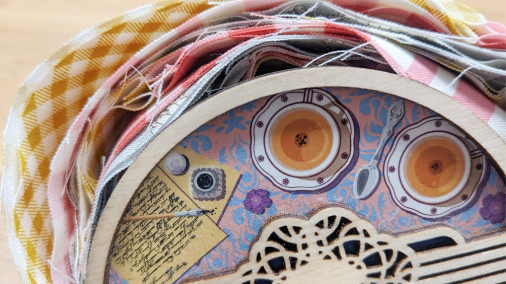 A close-up of layered colorful fabric strips next to a circular box lid decorated with painted teacups, saucers, a lace pattern, and a note on a blue background.
