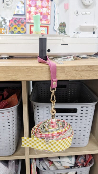 A strip of yellow gingham and floral fabric is rolled up and hanging from a gold clasp attached to a pink strap, which is secured to a wooden shelf. Gray plastic baskets and a sewing machine are visible in the background.