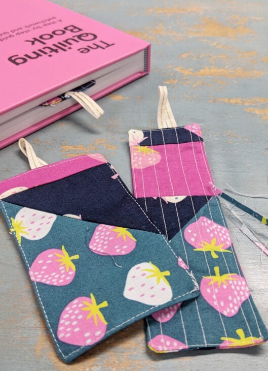 Fast & Easy DIY Fabric Bookmark | Use Up Leftover Quilt Blocks! 4 A pink sewing book and three DIY fabric bookmarks made from colorful patterns, including strawberries and stripes, lay on a blue-washed wooden surface.