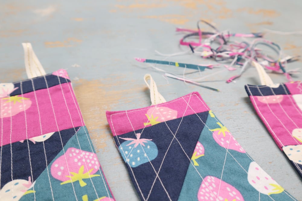 Fast & Easy DIY Fabric Bookmark | Use Up Leftover Quilt Blocks! 19 Three colorful quilted DIY fabric bookmarks with strawberry patterns and hanging loops are laid on a blue wooden surface. Scraps of trimmed fabric hint at easy sewing and creative quilt blocks in the background.
