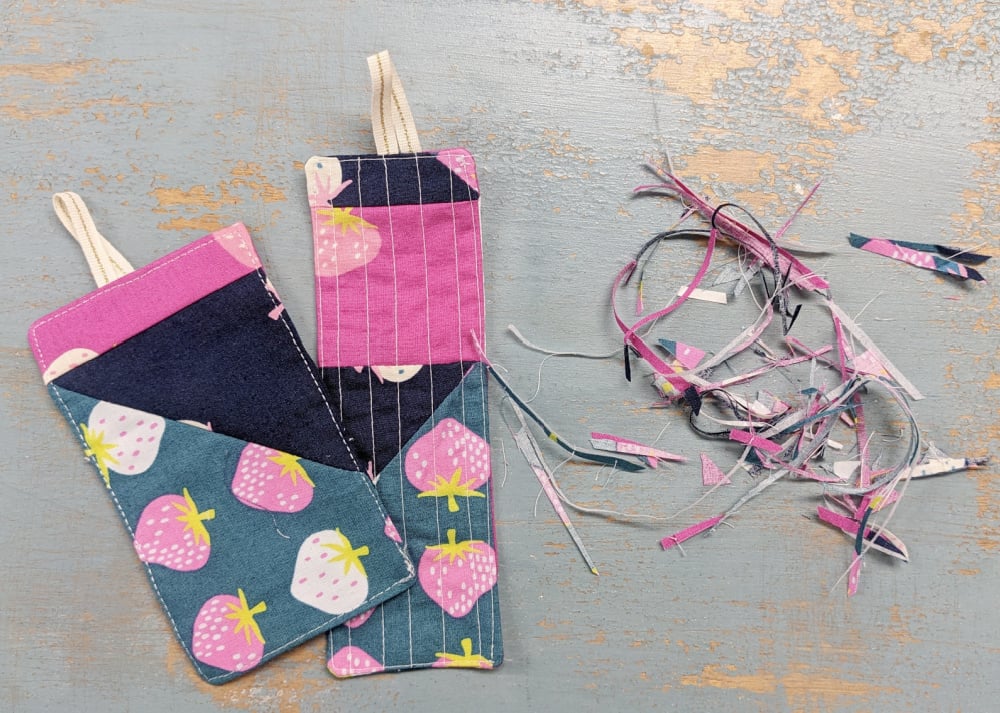 Fast & Easy DIY Fabric Bookmark | Use Up Leftover Quilt Blocks! 5 Two quilted DIY fabric bookmarks featuring pink, denim, and strawberry-patterned sections rest on a rustic wooden surface beside a pile of colorful leftover fabric trimmings.