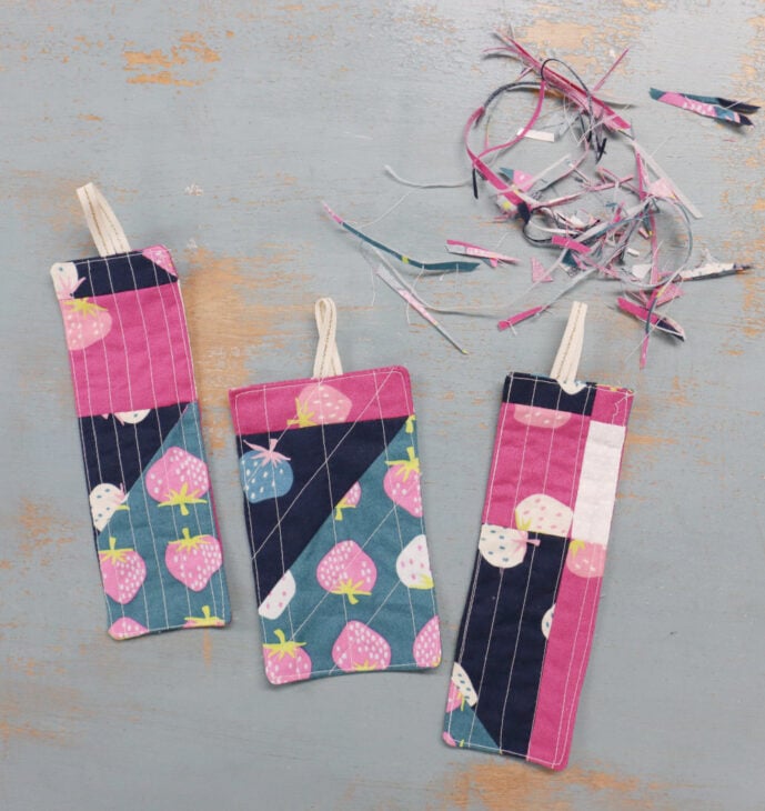 Fast & Easy DIY Fabric Bookmark | Use Up Leftover Quilt Blocks! 2 Three handmade DIY fabric bookmarks with pink, blue, and strawberry patterns lie on a distressed blue surface. Strips of fabric scraps are in the corner. Each bookmark has a small loop of ribbon at the top.