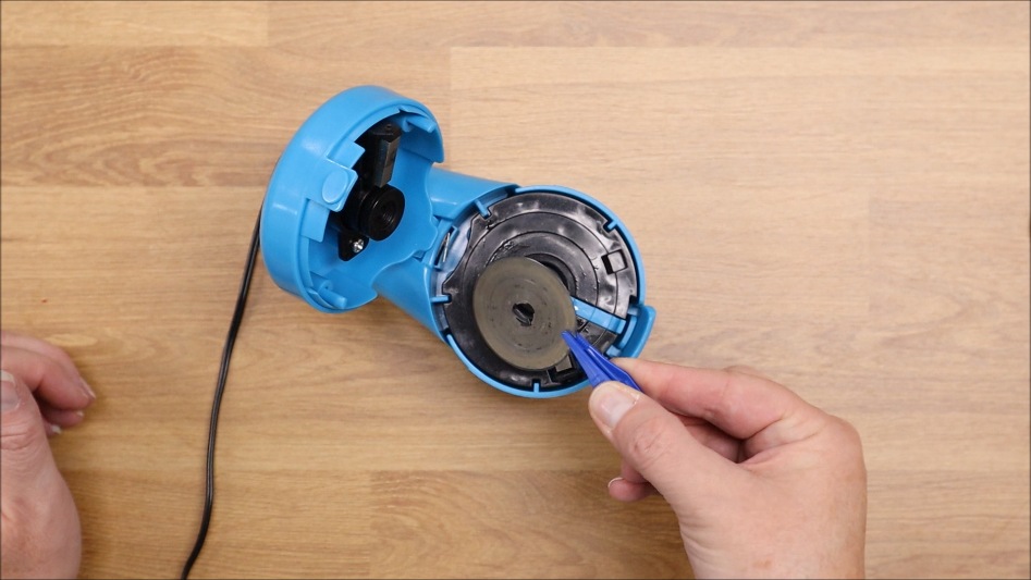 A hand holds a blue pen, pointing at the metal disc inside an open blue electric pencil sharpener on a wooden surface—perfect for a Blade Sharpener Tutorial or exploring how the TrueSharp rotary cutter blade sharpener works.