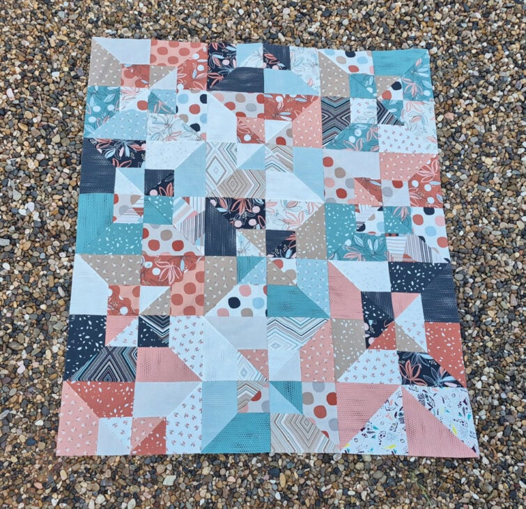 A colorful patchwork quilt top with geometric patterns in blue, peach, coral, white, and beige fabrics lies on a gravel surface—an inspiring creation that would be perfect for your next Virtual Quilt Retreat.