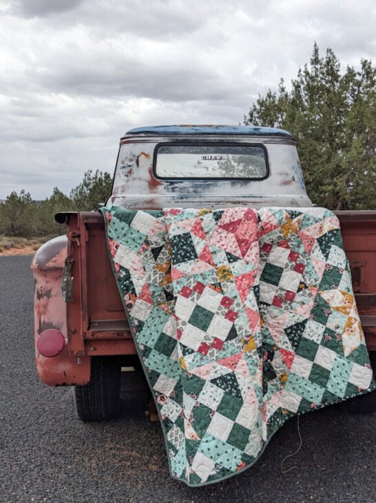Hidden Gems Block of the Month Finishing 3 A colorful patchwork quilt, showcasing expert quilting and quilt finishing, drapes over the tailgate of a rusty vintage red pickup parked on a gravel road, with trees and a cloudy sky in the background.