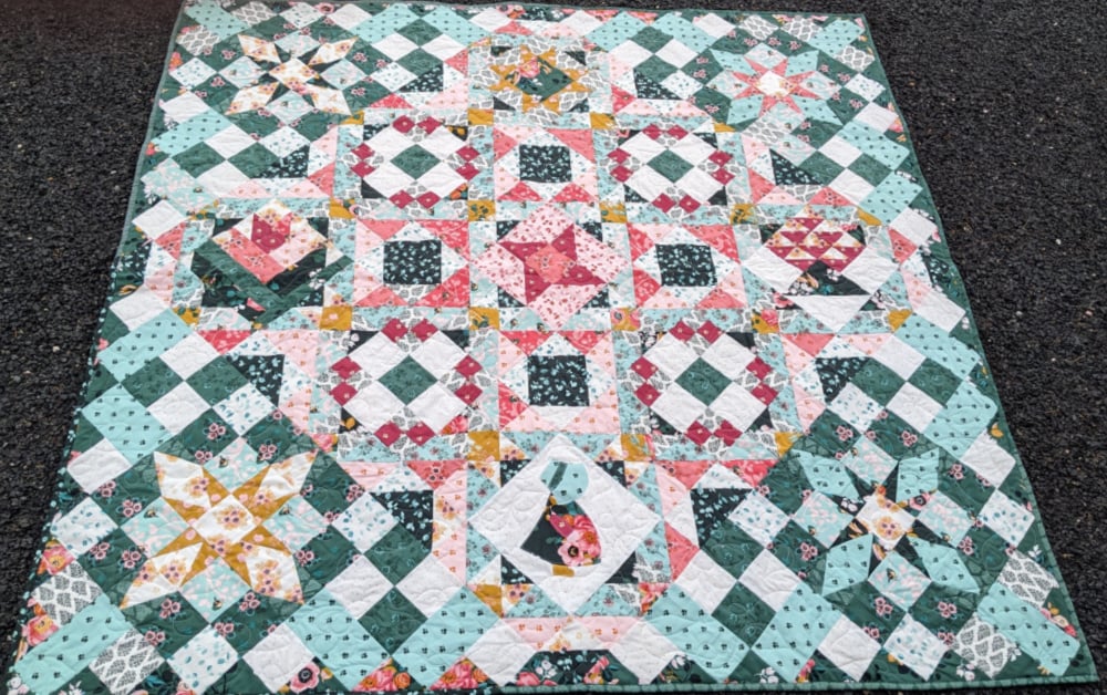 Hidden Gems Block of the Month Finishing 2 A colorful patchwork quilt from the Hidden Gems Block of the Month, with intricate geometric patterns in pink, teal, yellow, and white, laid out on a gravel surface. Diamonds, squares, and floral prints complete this beautifully finished quilt.
