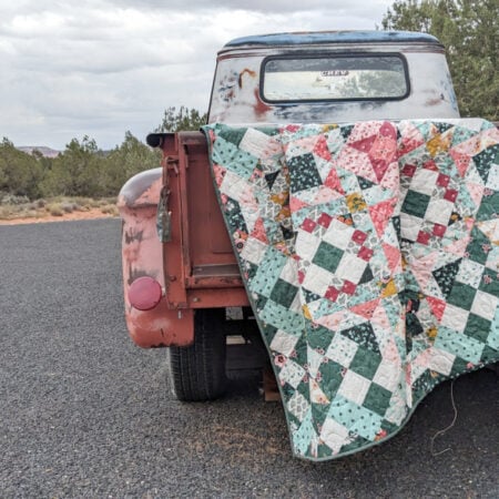 A weathered, rusty red pickup truck is parked on a paved road. An old, colorful patchwork quilt—perhaps part of a Hidden Gems Block of the Month quilting project—is draped over the open tailgate. Trees and cloudy sky fill the background.