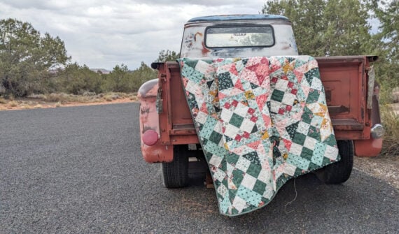 A weathered, rusty red pickup truck is parked on a paved road. An old, colorful patchwork quilt—perhaps part of a Hidden Gems Block of the Month quilting project—is draped over the open tailgate. Trees and cloudy sky fill the background.