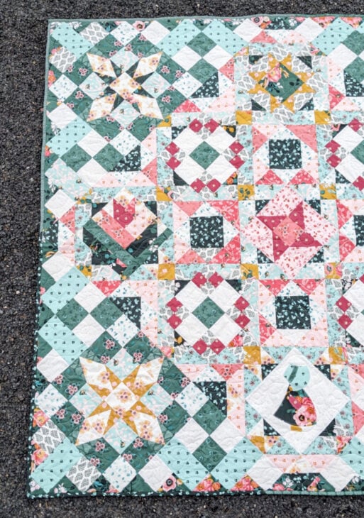 A handmade quilt featuring intricate patchwork in green, pink, white, and yellow floral patterns, with star, diamond, and heart shapes, reminiscent of the Hidden Gems Block of the Month, and a rabbit applique in the lower right corner, laid on a gravel surface.