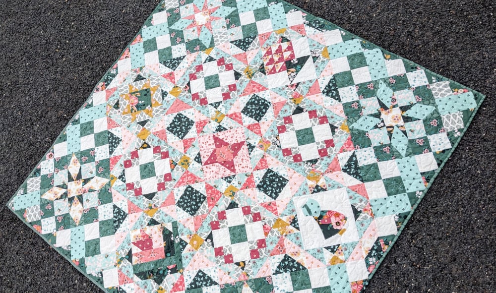 Hidden Gems Block of the Month Finishing 5 A colorful quilt with intricate geometric and star patterns in green, pink, white, and yellow, created for the Hidden Gems Block of the Month, displayed on a dark, textured surface.
