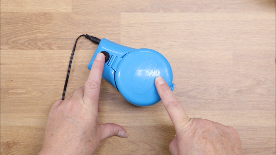 A person presses the button on a blue retractable dog leash with one hand while pointing to it with the other hand, next to a TrueSharp Rotary Cutter Blade Sharpener on a wooden surface.