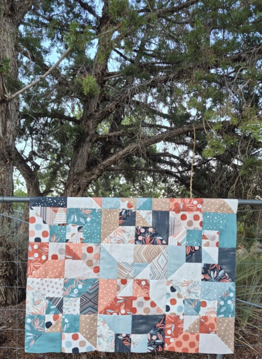 A colorful patchwork quilt with geometric patterns in blue, coral, cream, and brown hangs on a clothesline outdoors, with tall trees and green foliage in the background—a perfect scene to inspire your next Quilt Retreat.