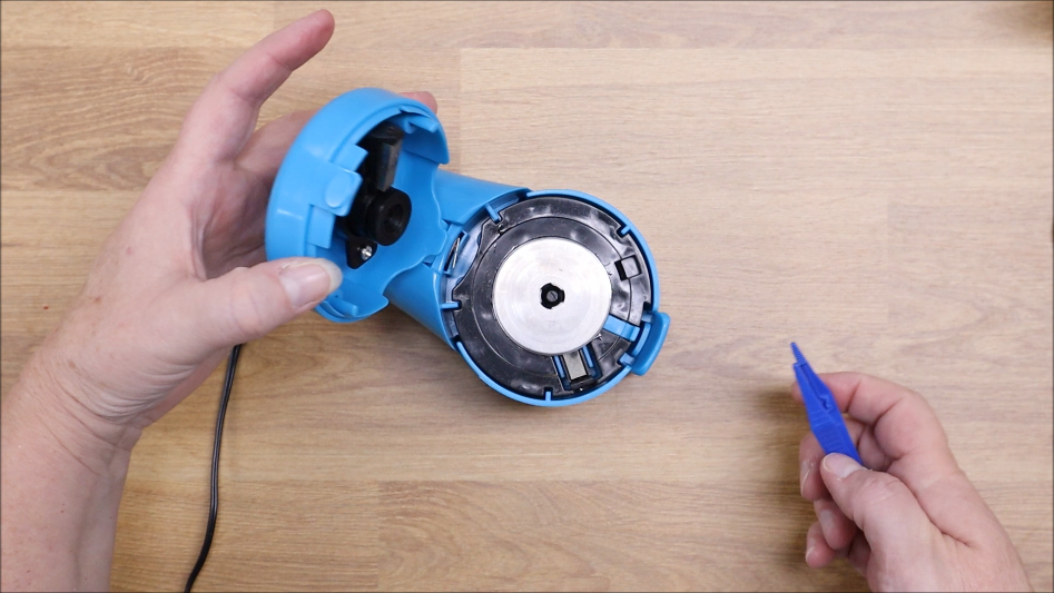 A person demonstrates a TrueSharp Rotary Cutter Blade Sharpener tutorial by holding the open blue appliance on a wooden surface, revealing its metal interior, while another hand uses a blue plastic tool.