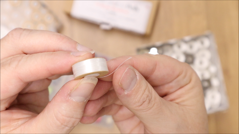 How to Use Pre-Wound Bobbins 1 A close-up of hands holding a white sewing thread spool, with a thin piece of thread extended. Boxes with pre-wound bobbins and additional spools are blurred in the background on a wooden surface.