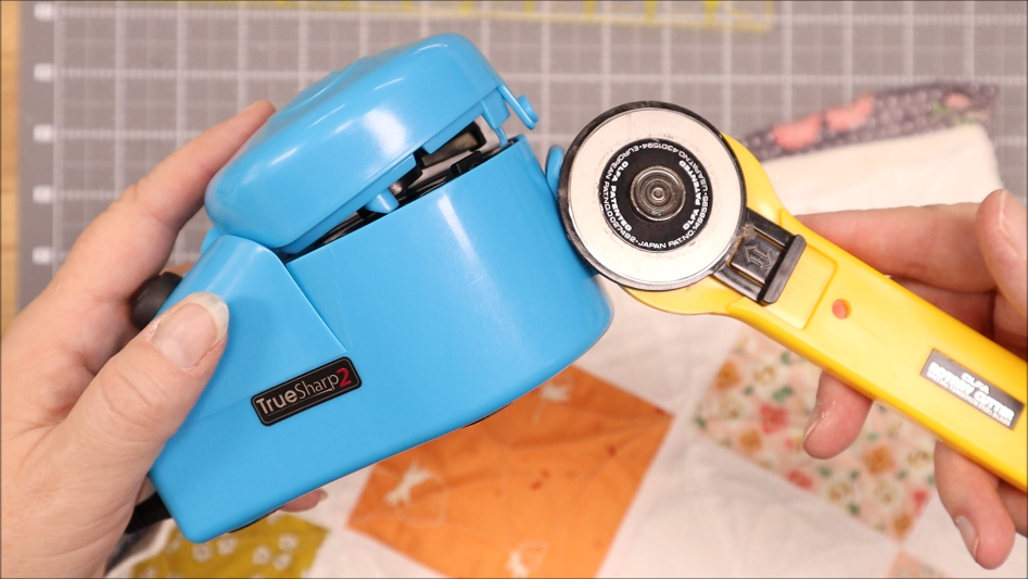 TrueSharp Rotary Cutter Blade Sharpener Tutorial 32 A hand holds a blue TrueSharp Rotary Cutter Blade Sharpener next to a yellow rotary cutter, with a cutting mat and quilted fabric squares in the background.