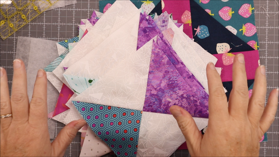 Fast & Easy DIY Fabric Bookmark | Use Up Leftover Quilt Blocks! 6 Two hands hold a stack of colorful quilt blocks on a gridded cutting mat. The fabric pieces, perfect for easy sewing projects like a DIY fabric bookmark, feature purple batik, blue hexagons, strawberries, and other vibrant designs.