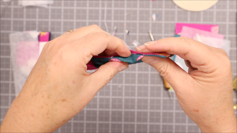 Fast & Easy DIY Fabric Bookmark | Use Up Leftover Quilt Blocks! 16 Close-up of two hands pinching colorful leftover quilt blocks together over a gray cutting mat with a grid pattern and scattered craft materials—perfect inspiration for easy fabric crafts.