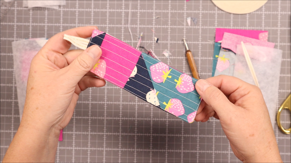 Fast & Easy DIY Fabric Bookmark | Use Up Leftover Quilt Blocks! 17 Two hands hold a rectangular DIY fabric bookmark, decorated with pink and blue stripes and strawberry patterns, on a crafting mat scattered with sewing tools and leftover quilt blocks in the background.