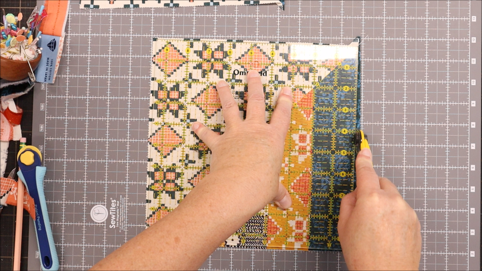 A person uses a rotary cutter and a clear measuring ruler to trim a colorful patchwork layer cake quilt square on a gray gridded cutting mat. Various sewing tools are visible, making the workspace perfect for projects that are simple to sew.