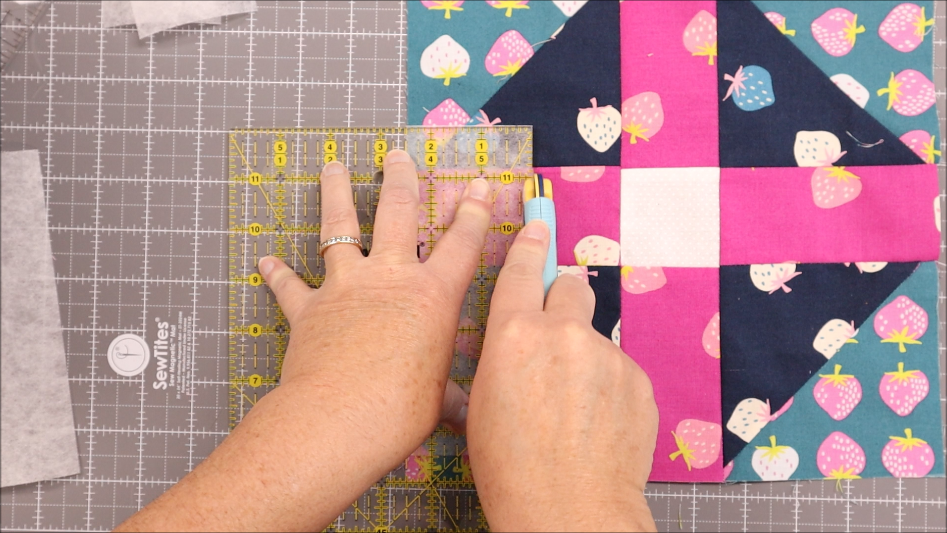 Fast & Easy DIY Fabric Bookmark | Use Up Leftover Quilt Blocks! 9 A person uses a rotary cutter and clear ruler to cut fabric pieces on a gridded cutting mat—perfect for creating quilt blocks or an easy sewing project like a DIY fabric bookmark. A colorful quilt block with strawberry patterns is visible to the right.