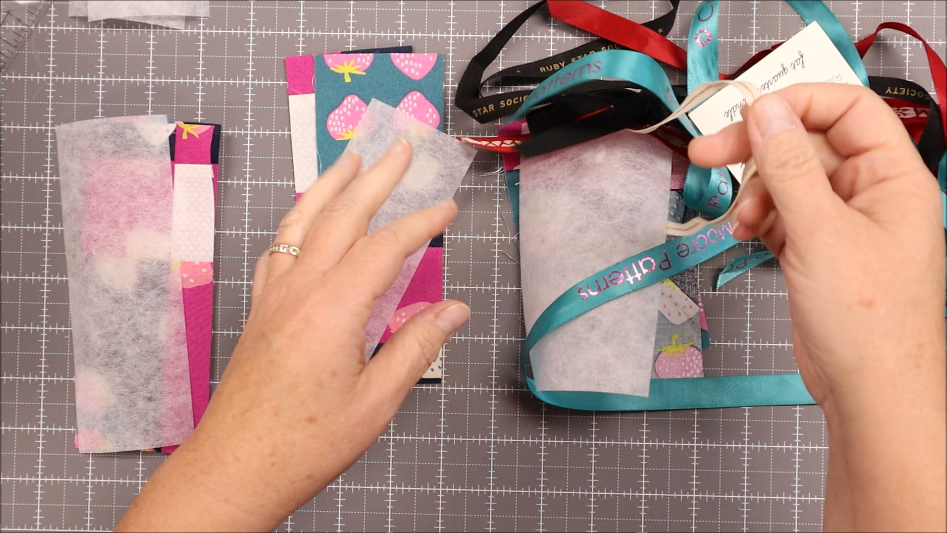 Fast & Easy DIY Fabric Bookmark | Use Up Leftover Quilt Blocks! 10 A person arranges colorful ribbon and patterned fabric pieces on a gridded cutting mat, preparing materials for an easy bookmark craft. One hand holds a ribbon, while the other adjusts a piece of white tissue paper.