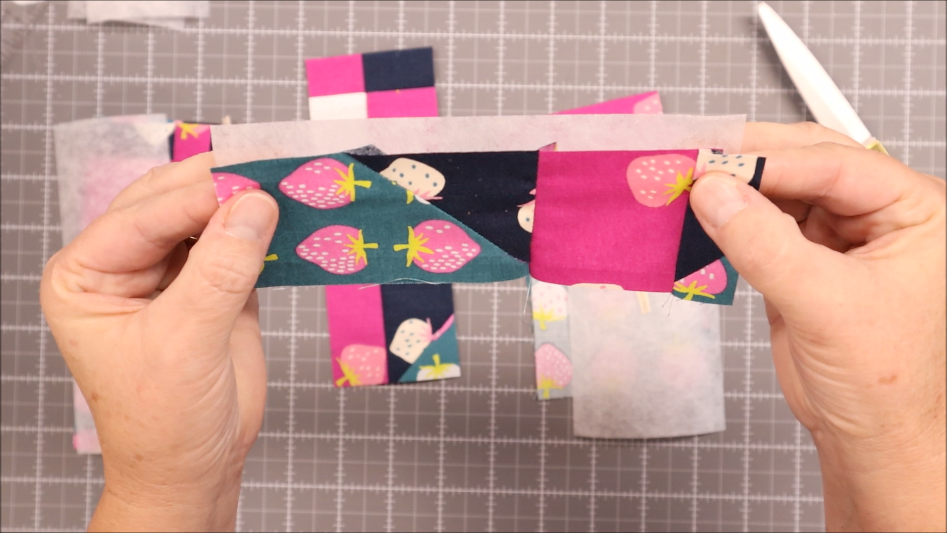 Fast & Easy DIY Fabric Bookmark | Use Up Leftover Quilt Blocks! 11 Two hands hold a DIY fabric bookmark pieced from colorful strawberry-patterned and pink leftover fabric strips, positioned over a cutting mat with other quilt blocks, scissors, and white interfacing in the background.
