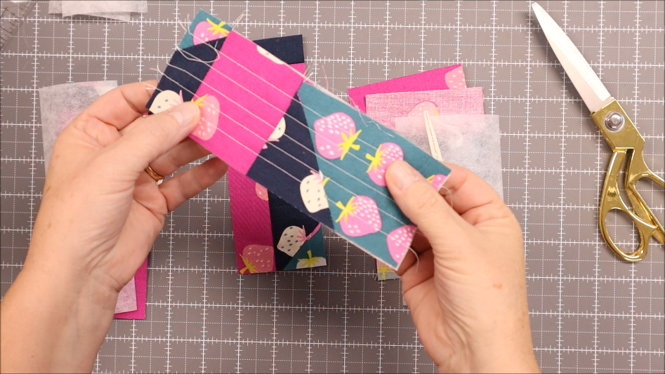 Fast & Easy DIY Fabric Bookmark | Use Up Leftover Quilt Blocks! 12 A person holds a piece of fabric with pink, blue, and strawberry patterns, showing stitched lines—perfect for easy fabric crafts like a DIY fabric bookmark. Scissors, leftover quilt blocks, and a gridded cutting mat are visible in the background.
