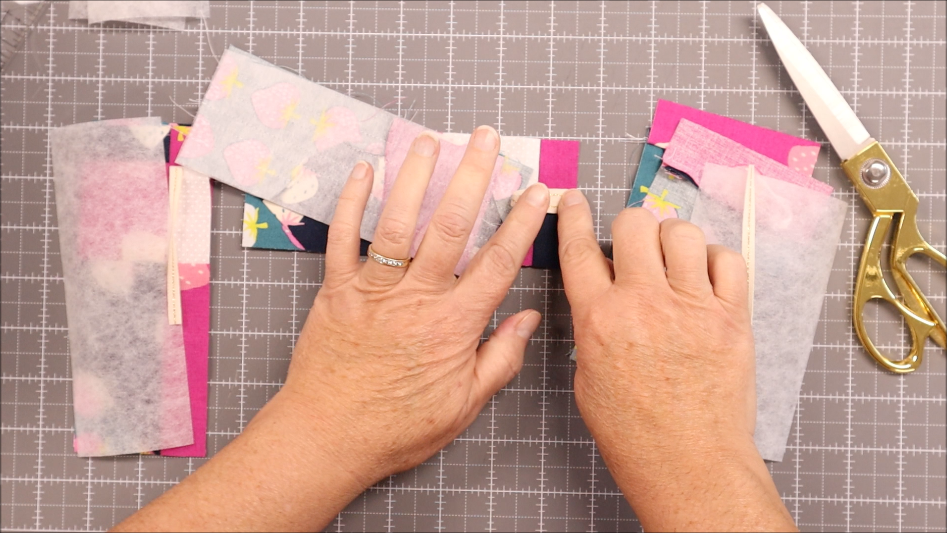 Fast & Easy DIY Fabric Bookmark | Use Up Leftover Quilt Blocks! 13 Hands arranging strips of colorful fabric and interfacing—perhaps from leftover quilt blocks—on a gridded cutting mat, with a pair of gold scissors nearby, ready to craft an easy bookmark.