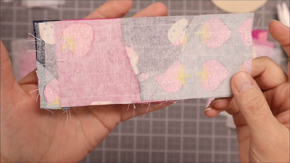 Fast & Easy DIY Fabric Bookmark | Use Up Leftover Quilt Blocks! 14 A person holds a rectangular piece of fabric with light pink and blue patterns, showing stitched lines across it. Made from leftover quilt blocks, the fabric appears to be sewn quilt pieces, viewed from the back side.