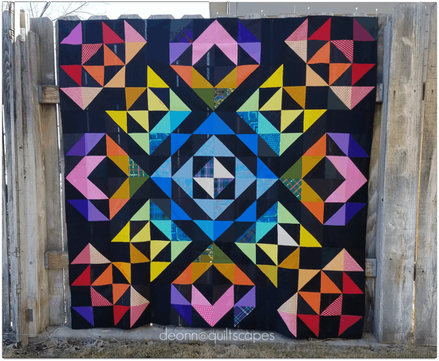 A colorful geometric quilt with vibrant triangles and squares arranged in a symmetrical pattern on a black background hangs on a wooden fence outdoors, showcasing edges that wow and finish it with flair.