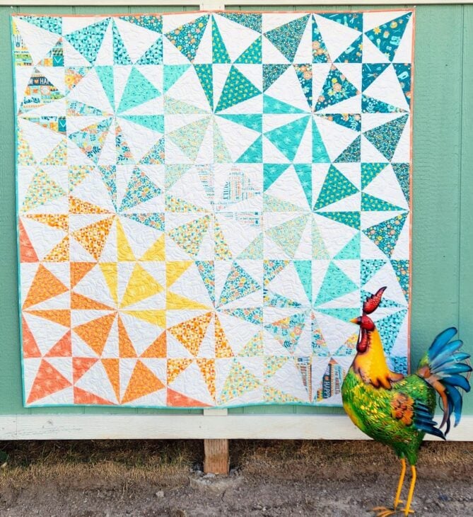 A colorful quilt with triangles in yellow, orange, teal, and white patterns hangs on a green wall. Its edges showcase binding basics to finish it with flair. In front is a vibrant metal rooster sculpture with blue, green, yellow, and red details.