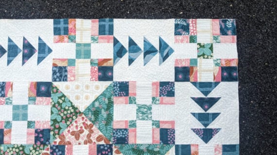 A colorful quilt with geometric patterns in shades of pink, green, blue, and white is displayed on a dark speckled surface. The quilt features triangles, squares, and floral fabric designs.