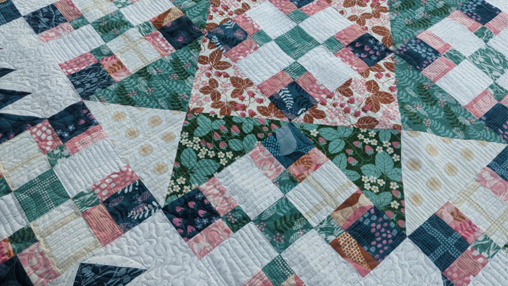A close-up of a colorful patchwork quilt featuring various floral, plaid, and polka dot fabric patterns in pink, green, blue, and white tones with intricate stitched detailing.