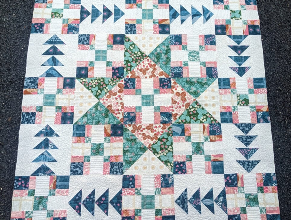 Crosswind Mystery Quilt Reveal 34 A handmade quilt featuring a large central star pattern in floral and geometric fabrics, surrounded by patchwork squares and flying geese triangles in shades of green, pink, blue, and white. The quilt lies on a dark textured surface.
