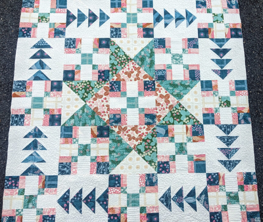 A colorful quilt featuring geometric patterns with stars, squares, and flying geese shapes in shades of blue, green, pink, and cream, arranged in a symmetrical, intricate design.
