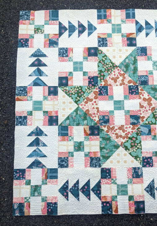 A colorful patchwork quilt featuring geometric patterns, including squares, triangles, and flying geese shapes, in shades of blue, green, pink, white, and tan, laid out on a dark textured surface.
