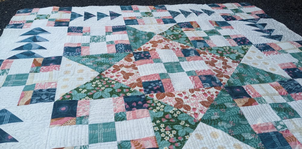 A colorful patchwork quilt features a large central star pattern, smaller square patterns, and flying geese shapes, using floral and geometric fabrics in green, pink, blue, and white tones. The quilt is spread out on the ground.
